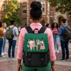 May include: Black backpack with a green flap featuring a graphic of several people in pink and white outfits. The backpack has black straps and buckles. The person wearing the backpack is standing in front of a brick building with other people in the background.