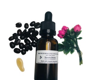 May include: A brown glass bottle with a dropper top filled with a clear liquid. The bottle is labeled "Brewed Rose Eye Concentrate with Raw Cacao" and "Navoms Beauty". The bottle is surrounded by coffee beans and a pink flower.