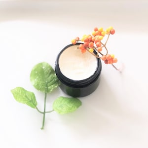 May include: A black jar filled with a white cream, with a sprig of orange and yellow berries on top. The jar is sitting on a white surface with a green leaf.