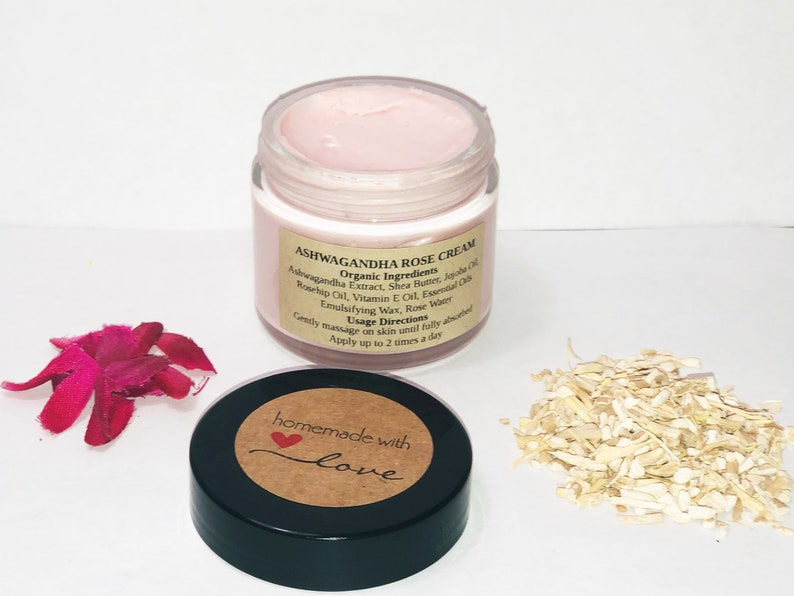 May include: A small glass jar with a pink cream inside. The jar has a brown label with the text "Ashwagandha Rose Cream" and a list of ingredients. The label also says "Organic Ingredients" and "Usage Directions". The jar is sitting on a white surface with a pink flower petal and a pile of shredded wood.