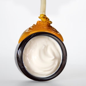 May include: A close-up of a white cream in a black jar with a golden liquid dripping over the top.