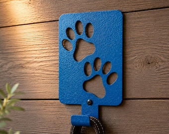 May include: A blue metal wall hook featuring two cutout paw prints. The rectangular plate has a textured finish and is secured with a single screw. A brown fabric leash with a metal swivel clasp hangs from the hook, which is mounted on a wall.