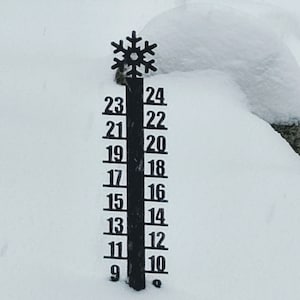May include: A black metal snow depth gauge with a snowflake design at the top. The gauge has numbers from 9 to 24.