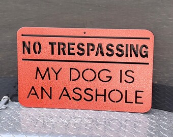 May include: A rectangular orange metal sign with rounded corners and a textured finish. The sign features black cut-out lettering that reads NO TRESPASSING MY DOG IS AN ASSHOLE. There is a small circular hole at the top center for mounting purposes.