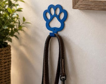 May include: A blue metal wall hook shaped like a paw print, mounted on a wall. A brown leather dog leash with a black metal clasp hangs from the hook. The hook features a cutout design in the shape of a paw, providing a sturdy point for hanging.