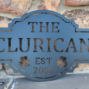 May include: A black metal sign with a rounded top and a decorative border. The sign reads "The Clurican Est 2001" with two shamrock designs on either side of the "Est 2001" text.