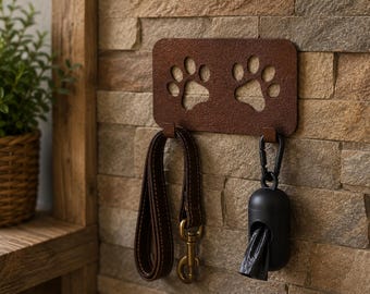 May include: A rectangular brown metal wall hook featuring two cut-out paw prints. The hook holds a dark brown leather leash with a brass-colored metal clip and a black plastic waste bag dispenser with a black carabiner clip attached to the hooks.