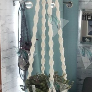 May include: A cream-colored macrame plant hanger with a white pot holding green plants. The hanger has a series of wooden beads and a tassel at the bottom. The background includes a wooden wall and a gray couch.