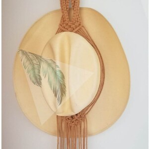 May include: A tan macrame hat holder with a wooden ring and hanging fringe. A light yellow hat with a green and white leaf design is displayed in the holder. The hat holder is mounted on a white wall.