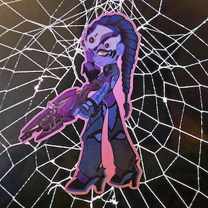May include: A sticker featuring a cartoon illustration of a character in a purple and blue outfit, holding a purple weapon. The character has a long blue braid and a mask with red accents. The background is a black surface with a white spiderweb design.