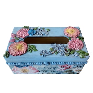May include: A light blue rectangular tissue box decorated with floral designs. The box has raised pink, yellow, and blue flowers, with green leaves. The sides of the box feature a floral pattern and script.