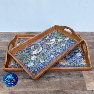 May include: Two rectangular wooden serving trays with cutout handles, featuring a dark blue background with a repeating pattern of light-colored birds, green foliage, white flowers, and small red berries printed on the base of each tray.