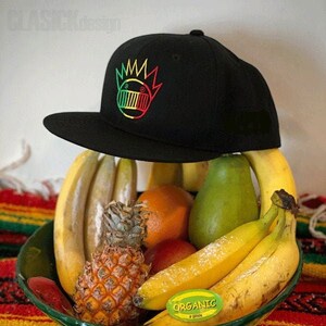 May include: A black and green baseball cap with a graphic of a cartoon face with a crown. The cap is sitting on a bowl of fruit, including bananas, a pineapple, oranges, and a pear. The sticker on the banana reads "Organic Brown".