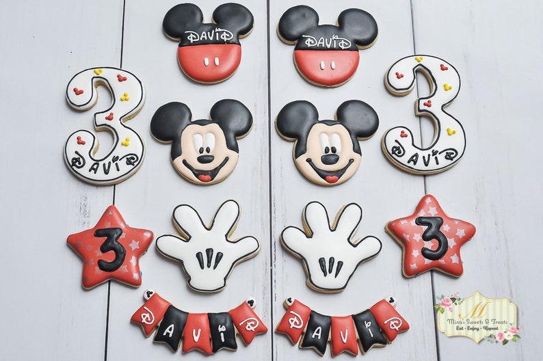 May include: A set of 12 Mickey Mouse themed sugar cookies decorated with red, black, white, and yellow icing. The cookies feature Mickey Mouse's face, his signature gloves, a star with the number 3, and the number 3 with Mickey Mouse ears. The cookies also include a banner with the name "David" and a Mickey Mouse head with the name "David".