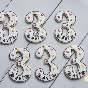 May include: Six white sugar cookies decorated with red and yellow sprinkles. Each cookie is shaped like the number "3" and has the name "DAVIR" written in black icing.