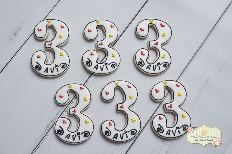 May include: Six white sugar cookies decorated with red and yellow sprinkles. Each cookie is shaped like the number "3" and has the name "DAVIR" written in black icing.