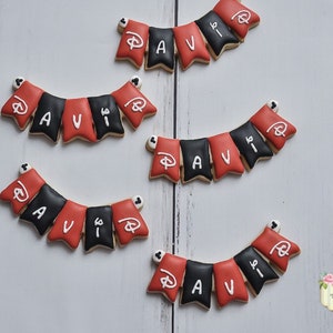 May include: Five red and black cookies decorated with white icing to spell out the name "AVI" with a Mickey Mouse head on each cookie. The cookies are arranged in a banner shape.
