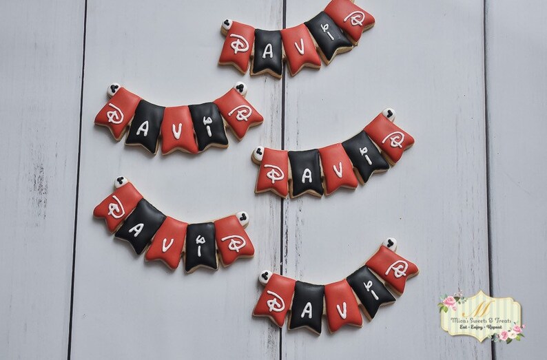 May include: Five red and black cookies decorated with white icing to spell out the name "AVI" with a Mickey Mouse head on each cookie. The cookies are arranged in a banner shape.