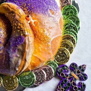 May include: A close-up of a King Cake, a traditional pastry with purple, green, and gold sprinkles. The cake is decorated with a small plastic baby and surrounded by gold and green coin-shaped candies, and a fleur-de-lis.