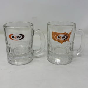 May include: Two clear glass A&W mugs with handles. One mug features the A&W logo in orange and brown. The other mug has the A&W logo inside an outline of the United States in orange and brown.