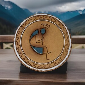 May include: A round, ceramic Kokopelli art piece. The central image features a stylized figure playing a flute, with blue and brown tones. The outer edge has a geometric pattern and a white border. The background shows a mountain range.