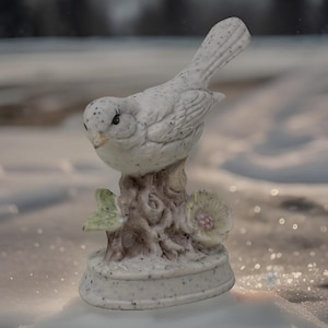 May include: A ceramic bird figurine with a speckled white finish perched on a textured tree stump base. The base features a small green leaf and a pale flower with a pink center. The bird has detailed wing feathers and a pointed beak.