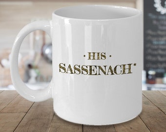 May include: White ceramic mug with the text "HIS SASSENACH" in gold glitter lettering. The mug has a standard handle and a smooth, glossy finish. The design is centered on the front of the mug.