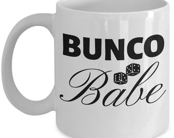 May include: White ceramic mug with the text "BUNCO Babe" in black. The word "BUNCO" is in a bold, sans-serif font, and "Babe" is in a cursive script. Two dice are between the words.
