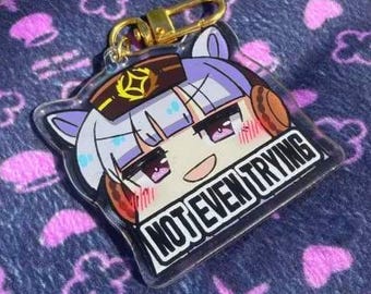 Puede incluir: Un llavero acrílico transparente con un personaje de estilo anime y el texto "NOT EVEN TRYING". El llavero está sujeto a un cierre dorado. Descansa sobre una tela azul marino estampada con flores y cruces rosas.