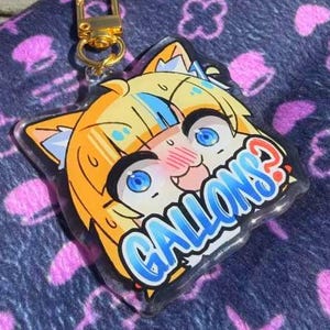 May include: A clear acrylic keychain featuring a cartoon cat girl with orange hair and blue eyes. The keychain has a gold-colored clasp and the word "GALLONS?" in blue and white text. The background is a dark blue fabric with pink patterns.