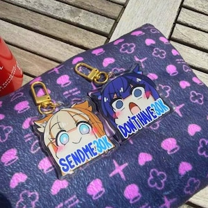 May include: Two anime-style keychains with gold-colored clasps. One keychain features a character with orange hair and the text "SEND ME 30K". The other keychain has a blue-haired character and the text "DON'T HAVE 30K". They rest on a dark blue patterned surface.