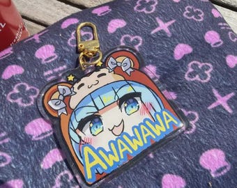 Puede incluir: Un llavero de acrílico transparente con un personaje de dibujos animados con cabello y ojos azules, y la palabra "AWAWAWA" en amarillo. El llavero tiene un cierre dorado y está sobre una superficie azul oscuro estampada con diseños florales y cruces rosas.
