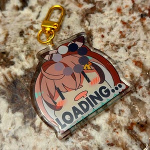 Puede incluir: Un llavero de acrílico transparente con un personaje de anime de pelo rosa y un atuendo azul y blanco. El personaje está mirando hacia abajo con una expresión de sorpresa y el texto "LOADING..." está impreso en el llavero.