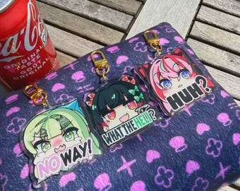 Puede incluir: Tres llaveros de estilo anime con cierres dorados. Cada llavero presenta un personaje diferente con ojos grandes y un bocadillo. Los bocadillos dicen "NO WAY!", "WHAT THE HELL?" y "HUH?". Los llaveros descansan sobre una superficie morada estampada.
