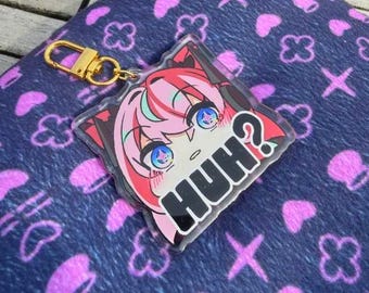 Puede incluir: Un llavero acrílico transparente con un personaje de anime de pelo rosa, ojos azules y la palabra "HUH?" en negro. Tiene un cierre dorado y está sobre una tela azul marino con patrones rosas.