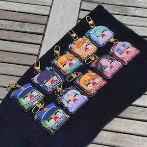 May include: A collection of anime-style acrylic keychains with gold-colored clasps. Each keychain features a colorful character face with the word "YOHHHHH!" in a speech bubble. The keychains are displayed on a black surface.
