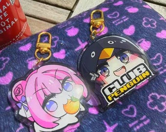 Puede incluir: Dos llaveros de estilo anime con cierres dorados. Uno presenta un personaje de pelo rosa sosteniendo una fruta, y el otro tiene un diseño de pingüino con el texto "CLUB PENGUIN". Ambos están sobre una superficie azul estampada.
