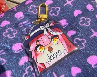 Puede incluir: Un llavero acrílico transparente con un personaje de anime de pelo rosa con un sombrero y la palabra "doom". El llavero tiene un cierre dorado y está sobre un fondo azul estampado.