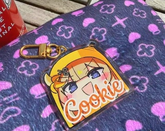 Puede incluir: Un llavero acrílico transparente con un personaje de dibujos animados con pelo amarillo y ojos azules. La palabra "Cookie" está escrita en naranja. El llavero tiene un cierre dorado y está sobre una tela azul estampada.
