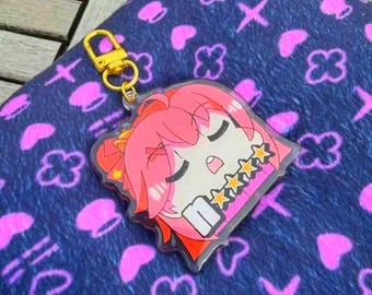 Puede incluir: Un llavero acrílico transparente con un personaje de anime de pelo rosa con una diadema adornada con estrellas. El llavero tiene un cierre dorado y está sobre un fondo azul estampado.