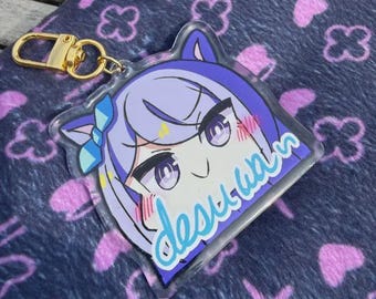 Puede incluir: Un llavero acrílico transparente con un personaje de anime con cabello morado y un lazo azul. El llavero tiene un cierre dorado y el texto "desu wa" en azul. El fondo es una tela azul oscuro con patrones florales rosas.