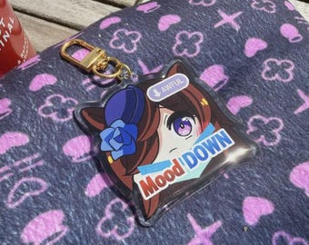 Puede incluir: Un llavero acrílico transparente con un diseño de personaje de anime de dibujos animados. El llavero tiene un cierre dorado y presenta el texto "Mood Down" y "Awful". El fondo es una tela azul oscuro con patrones florales rosas.