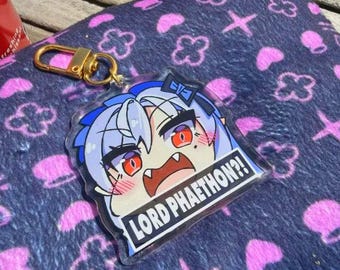 Puede incluir: Un llavero acrílico transparente con un personaje de estilo anime con cabello azul y ojos rojos. El llavero tiene un cierre dorado y el texto "LORD PHAETHON?!" en un rectángulo blanco.