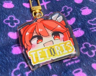 Puede incluir: Un llavero acrílico transparente con un personaje de anime de pelo y ojos rojos. El llavero tiene una pancarta amarilla con el texto "TETORIS". Tiene un cierre dorado.