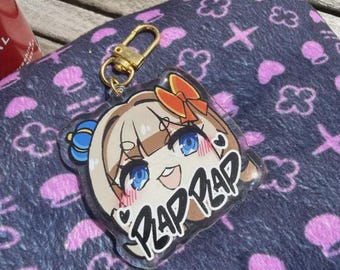 Puede incluir: Un llavero acrílico transparente con un personaje de estilo anime con ojos azules, un lazo rojo y el texto "PLAP PLAP". El llavero tiene un cierre dorado y está sobre una superficie estampada.