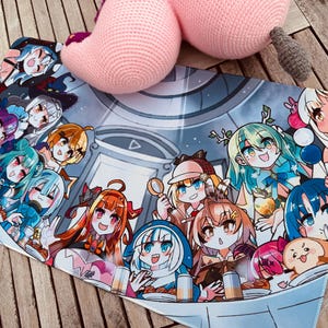 May include: A rectangular mousepad featuring a colorful anime-style illustration of multiple characters. The design includes a variety of characters with different hair colors and expressions. A pink crocheted object rests on the mousepad.
