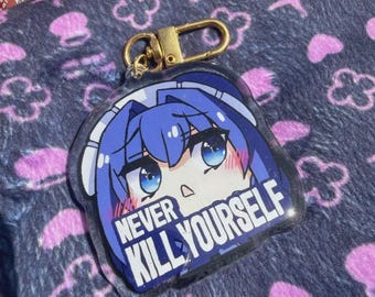 Puede incluir: Un llavero acrílico transparente con un personaje de estilo anime con cabello y ojos azules. El llavero tiene un cierre dorado y el texto "NEVER KILL YOURSELF". El fondo es una tela azul oscuro con patrones florales y de cruces rosas.