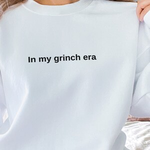 May include: White sweatshirt with the text "In my grinch era" printed on the front.