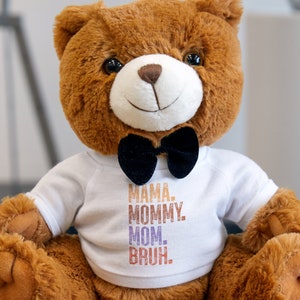 May include: A brown teddy bear wearing a white shirt with the text "MAMA. MOMMY. MOM. BRUH." printed on it. The bear is wearing a black bow tie.