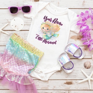 May include: A white bodysuit with a mermaid illustration and the text "Your Name the Little Mermaid". The bodysuit is paired with a rainbow iridescent mermaid tail skirt, purple flower lei, and white starfish.